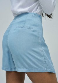 Bermudas de denim azul claro con una textura suave, que cuentan con un bolsillo trasero y un dobladillo cosido, cortadas por encima de la rodilla.