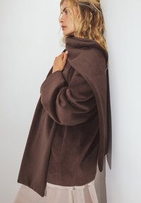 Pruun, ülemõõduline mantel pehmest kangast, millel on langev krae ja laiad varrukad, valge seina taustal. All on kerge, kihiline riietus.