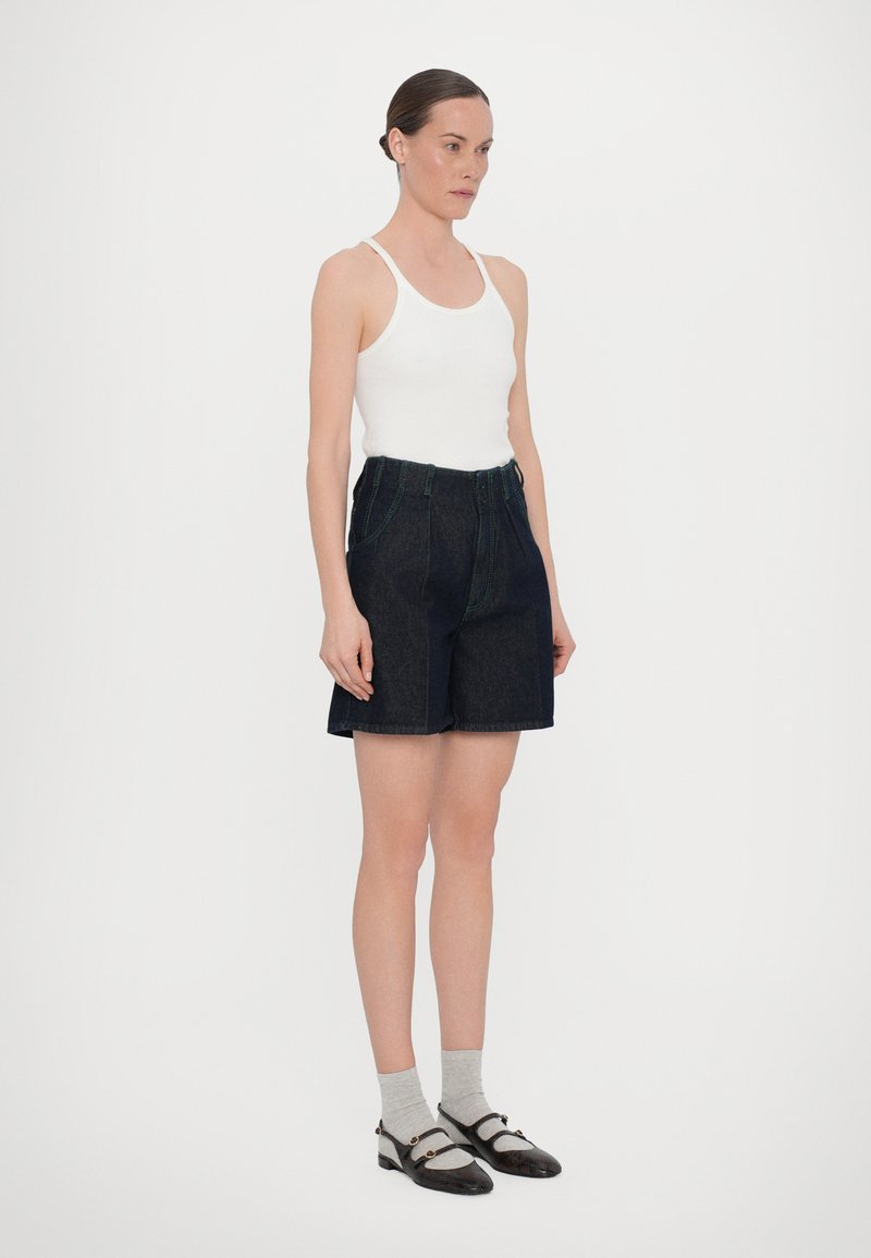 Dunkelblaue Denim-Shorts mit grünen Nähten, tailliertem Bund und vorderen Taschen. Kombiniert mit einem weißen Tanktop und grauen Socken, im Stand. 