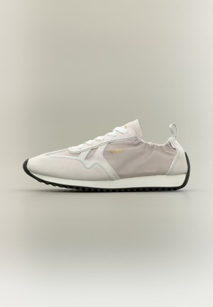 Baskets basses beige clair et grises avec lacets blancs, semelle noire, petit logo de marque doré, et languette élastique au talon sur fond uni.