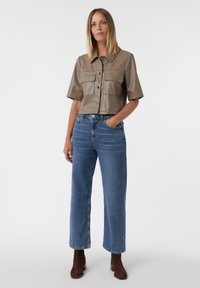 Abgeschnittenes, olivgrünes Lederhemd mit kurzen Ärmeln und zwei Brusttaschen, kombiniert mit hochtaillierten blauen Jeans im geraden Schnitt und braunen Stiefeletten.