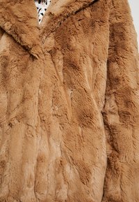 Brun fuskpälsjacka med en mjuk, strukturerad yta och ett vågmönster. Den har en krage med slag och ett varmt, fluffigt material.