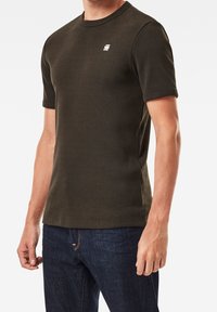 Olijfgroene, geribbelde t-shirt met korte mouwen en een ronde halslijn, voorzien van een klein logo op de borst, gecombineerd met donkerblauwe jeans.