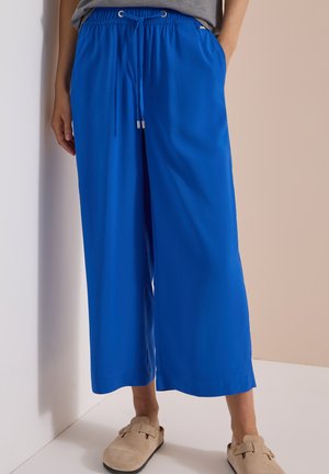 Person iført lyseblå bukser med vide ben, elastisk talje og snøre, kombineret med beige slip-on sko, stående op ad en væg.