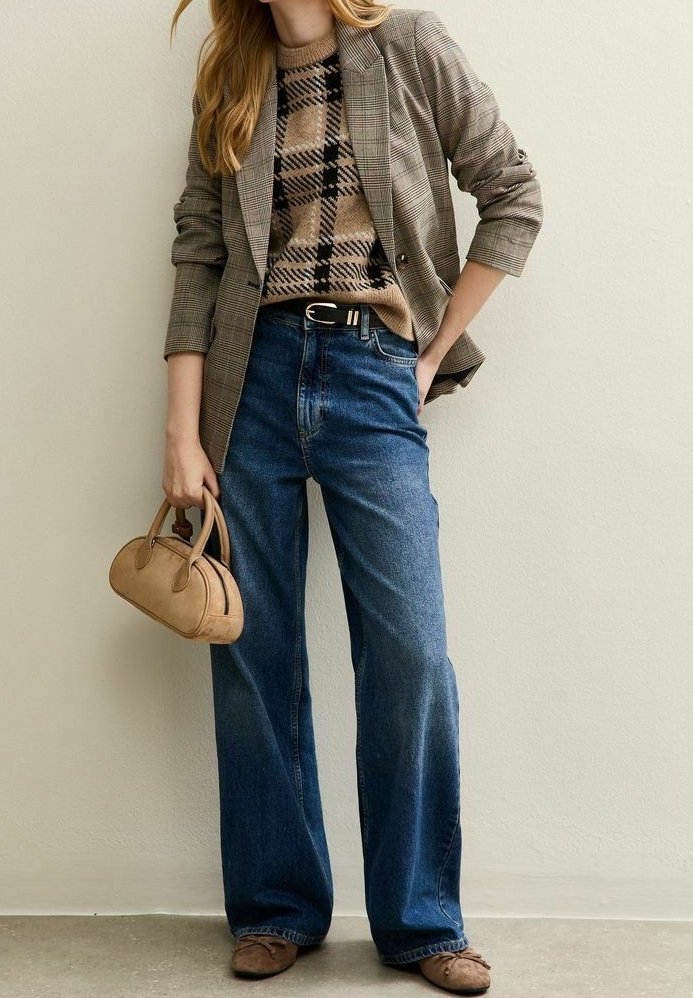 Femme portant un blazer à carreaux, un pull beige à motifs, un jean bleu à jambes larges, des chaussures marron, tenant un petit sac à main beige, debout contre un mur clair.