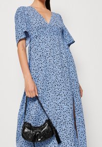Abito floreale blu con maniche corte e scollo a V, caratterizzato da un motivo di fiori neri. Si tiene in mano una piccola borsa nera con manico a catena.