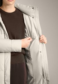 Geacă călduroasă gri deschis, cu o bluză tricotată texturată de culoare maro dedesubt. Geaca are capse și un finisaj neted, lucios.