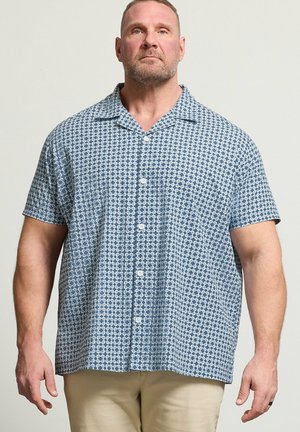 Mann mit kurzen Haaren und Bart, der ein blau gemustertes, kurzärmeliges Hemd und beige Hose trägt, steht vor einem einfarbigen Hintergrund.