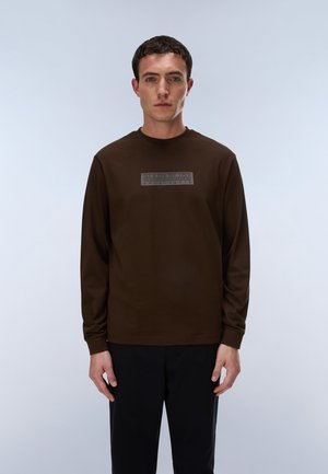 Homme portant une chemise à manches longues marron foncé avec un logo rectangulaire sur la poitrine, debout devant un fond clair uni.