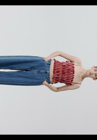 Femme portant un haut sans manches rayé rouge et blanc et un jean bleu taille haute, debout les mains dans les poches devant un fond uni.