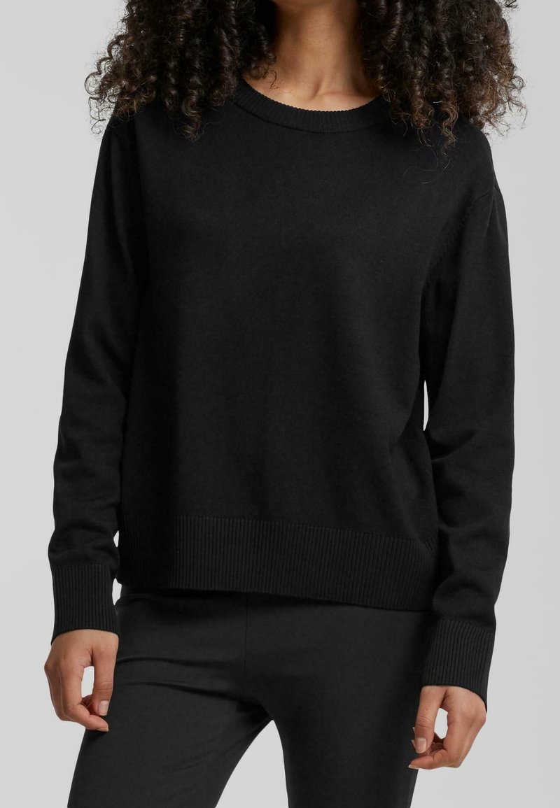 Personne aux cheveux bouclés portant un pull noir uni à manches longues et un pantalon noir sur un fond clair uni.