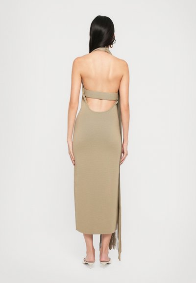 Woman with long dark hair wearing a backless beige midi dress with a halter neck and white heeled mules, standing against a plain white background.