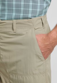Pantaloni beige con una texture liscia, dotati di una tasca laterale e dettagli di cucitura visibili. Indossati con una camicia a quadri blu e verde.