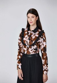 Blouse à manches longues avec un motif floral marron, dotée d'un col et de poignets noirs à volants, accompagnée d'une jupe plissée noire.