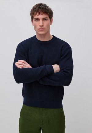 Junger Mann mit kurzen lockigen Haaren, der einen dunkelblauen Pullover und grüne Hosen trägt, steht mit verschränkten Armen vor einem schlichten hellen Hintergrund.