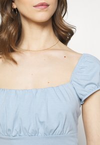 Mladá žena v světle modrém topu s nafouknutými rukávy, se zlatými kruhovými náušnicemi a tenkým zlatým náhrdelníkem, odhalující krk a horní část hrudníku s mateřskými znaménky.