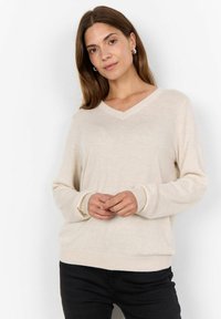 Maglione beige a collo a V con maniche lunghe, realizzato in materiale morbido. Presenta polsini e orlo a coste, vestibilità comoda. Indossato sopra pantaloni neri.