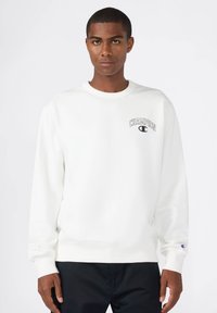 Biała bluz sweatshirtowa, z okrągłym dekoltem, z ribbowanymi mankietami i dołem, z czarnym logo "Champion" na piersi, miękka tekstura tkaniny. Ubrana czarna spódnica.