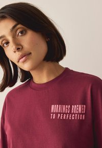 Burgundy pitkähihaisessa t-paidassa on vasemmassa rintakappaleessa vaaleanpunaista tekstiä, jossa lukee "MORNINGS BREWED TO PERFECTION". Pehmeää puuvillakangasta.