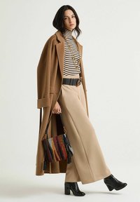 Pantaloni beige a gamba larga, cintura nera, dolcevita a righe, cappotto marrone, stivali neri con tacco e borsa a tracolla colorata con motivi e nappine.