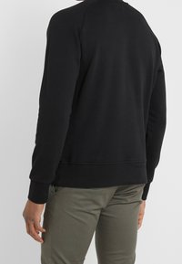 Sudadera negra con puños y dobladillo acanalados, con cuello redondo y un acabado de tela texturizada. Llevada sobre pantalones verdes, mostrando la vista trasera.
