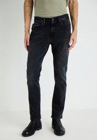 Juodų džinsų kelnės su siauru pjaustymu, penkiais kišenėmis ir subtiliomis siuvimo detalėmis. Dėvimos su šviesiai žalia marškinėliais ir juodais auliniais batais iki kulkšnies.