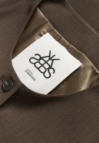 Rudas audinys drabužis su lygiu paviršiumi, turintis apvalią iškirptę, sagčių uždarymą ir priežiūros etiketę su juodu logotipo tekstu.