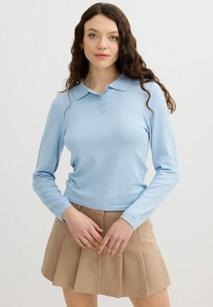 Mujer joven con el pelo largo y ondulado que lleva una camisa polo de manga larga azul claro y una falda plisada beige, de pie con los brazos cruzados.