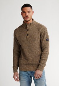 Man in een bruine, gestructureerde gebreide trui met een geknoopte kraag en lichtblauwe jeans, staand tegen een witte achtergrond.