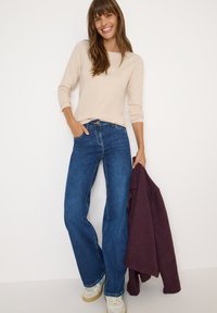 Beige lange mouw top gecombineerd met donkerblauwe wijde jeans. Lichtgekleurde sneakers en een gestructureerde bordeauxrode jas over één arm draped.
