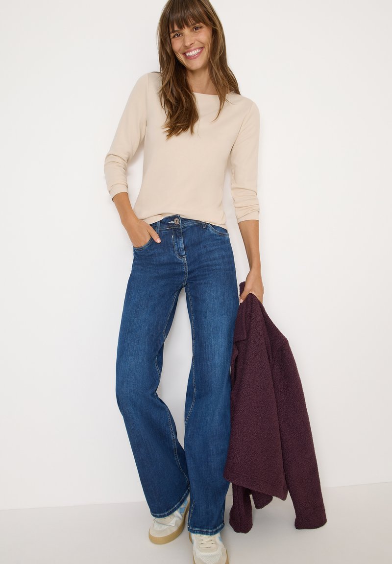 Beige lange mouw top gecombineerd met donkerblauwe wijde jeans. Lichtgekleurde sneakers en een gestructureerde bordeauxrode jas over één arm draped.