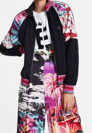 Veste noire avec manches à motif floral et poignets rayés roses sur une chemise blanche et un pantalon coloré à imprimé tropical.