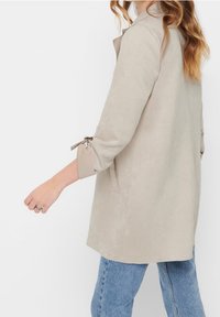 Kvinna i beige långärmad kappa med knytband vid ärmarna och blå jeans, sedd från sidan mot en enkel bakgrund.