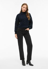 Granatowy sweter z dzianiny o splocie warkoczowym, krótki, z wysokim kołnierzem, zestawiony z czarnymi spodniami o wysokim stanie i botkami na kostkę, w prostym kroju nogawek.