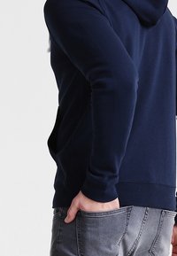 Sudadera con capucha de color azul marino, con un bolsillo tipo canguro, puños acanalados y una textura suave, combinada con jeans de mezclilla gris.