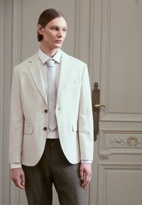 Light beige linen blazer with two buttons, paired with a pale shirt and light gray trousers. Simple design, tailored fit, textured fabric.