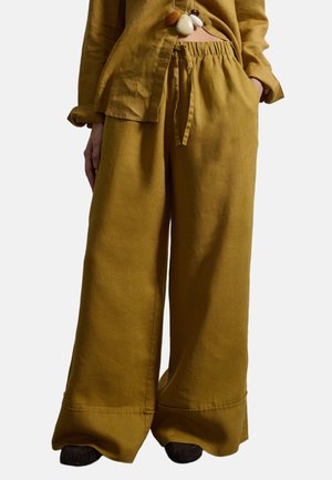 Frau trägt eine senfgelbe, weit geschnittene Hose mit Kordelzug und ein passendes, geknöpftes Hemd, dazu eine geschichtete Anhängerkette und schwarze Schuhe.