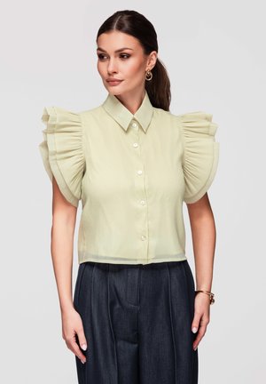 Kobieta nosząca bladoróżową bluzkę z falbankami na krótkich rękawach oraz ciemne plisowane spodnie z wysokim stanem, pozująca na jednolitym tle.