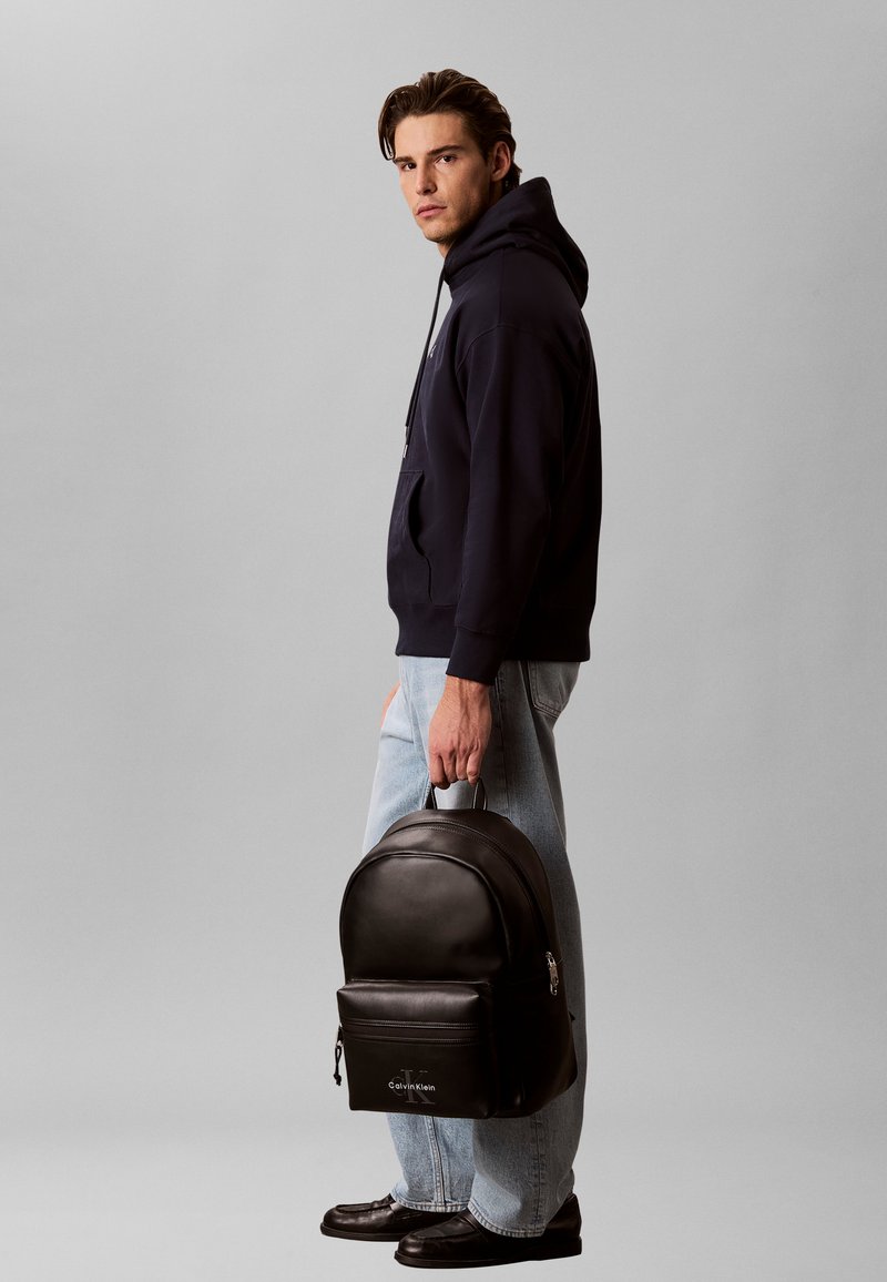 Homme en sweat à capuche noir et jean clair tenant un sac à dos noir Calvin Klein par la poignée, debout de profil sur fond gris.