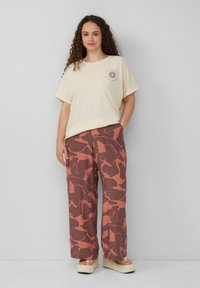 Cremfarbener T-Shirt mit kurzen Ärmeln und Sonnenmotiv auf der Brust, kombiniert mit weit geschnittenen Hosen mit einem abstrakten rost- und braunfarbenen Muster.