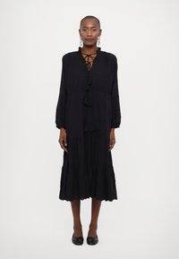 Vestido preto de mangas longas com decote com cordões e detalhes em franjas. Apresenta um corpete bordado e uma saia em camadas e fluida.