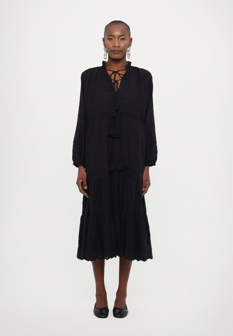 Vestido preto de mangas longas com decote com cordões e detalhes em franjas. Apresenta um corpete bordado e uma saia em camadas e fluida.