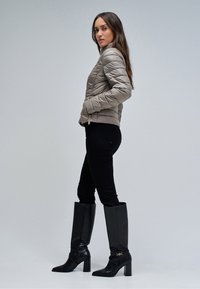 Vrouw staand zijwaarts, gekleed in een grijze gewatteerde jas, zwarte strakke broek en zwarte kniehoge laarzen met hak en gouden detail, tegen een effen achtergrond.