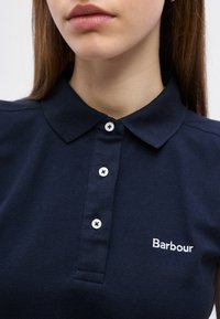 Γυναίκα που φοράει μπλε σκούρο πόλο πουκάμισο Barbour με τρία λευκά κουμπιά και γιακά, εμφανιζόμενη από τους ώμους έως τα χείλη.