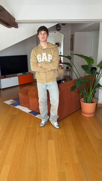 Camisola com fecho éclair bege com logótipo "GAP", jeans azul claro e sapatilhas prateadas. Sala com chão de madeira, sofá castanho e uma grande planta verde.