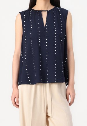 Sleeveless navy blouse with vertical white dot stripes and a front keyhole neckline, worn with loose beige pants.