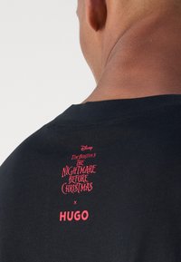 Musta puuvillapaita, jossa on punainen graafinen kuvasto, jossa lukee "Disney Tim Burtonin Painajainen ennen joulua" ja "HUGO"-logo takana kaula-aukon kohdalla.