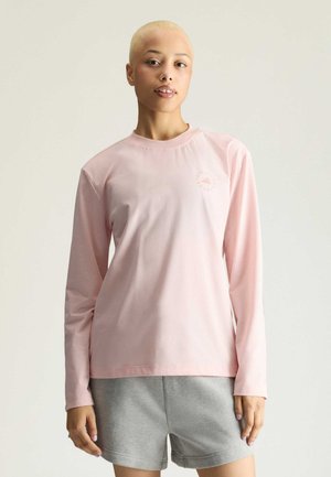 Ung person med kort blondt hår iført en lyserød langærmet skjorte og grå shorts, der står mod en ensfarvet lys baggrund.