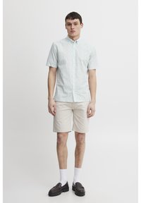 Koszula krótki rękaw, jasna zielona w wzory, z zapięciem na guziki, zestawiona z beżowymi szortami. Model nosi ciemne loafersy i białe skarpetki.