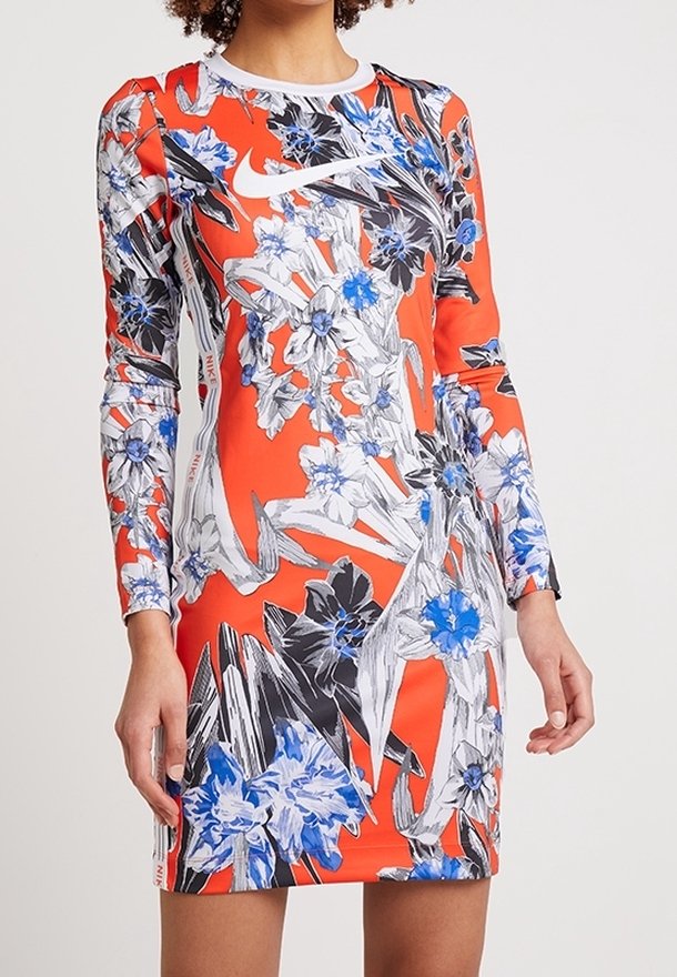 Donna che indossa un vestito aderente a maniche lunghe con uno sfondo arancione e motivi floreali blu, neri e bianchi, con un logo Nike bianco sul petto.
